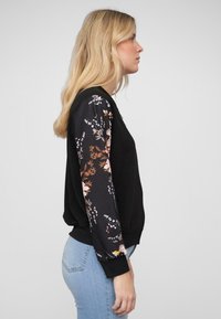 Schwarzes Sweatshirt mit blumenmusterbezogenen Ärmeln, gerippten Bündchen und Saum. Vorgestellt mit blauen Jeans, die einen lockeren Sitz und eine weiche Textur betonen.