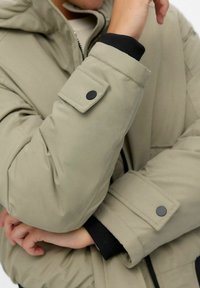 Pufová bunda v olivově zelené tkanině s černými akcenty. Má dlouhé rukávy se zapínáním na patentky a zip. Měkký, texturovaný povrch.