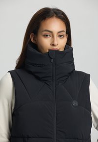 Gilet rembourré noir avec col montant, doté d'une fermeture éclair à l'avant, d'un design matelassé en diagonale et d'un logo circulaire sur la zone gauche de la poitrine.
