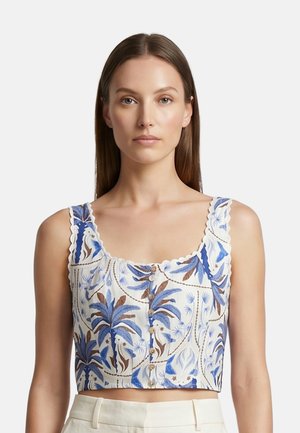 Vrouw draagt een mouwloos crop top met blauwe en bruine tropische bladpatronen en houten knopen, gecombineerd met lichte broek.