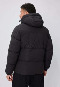 Doudoune noire avec capuche, présentant un design matelassé, des poignets élastiques et une coupe décontractée. Le matériau semble lisse et rembourré.