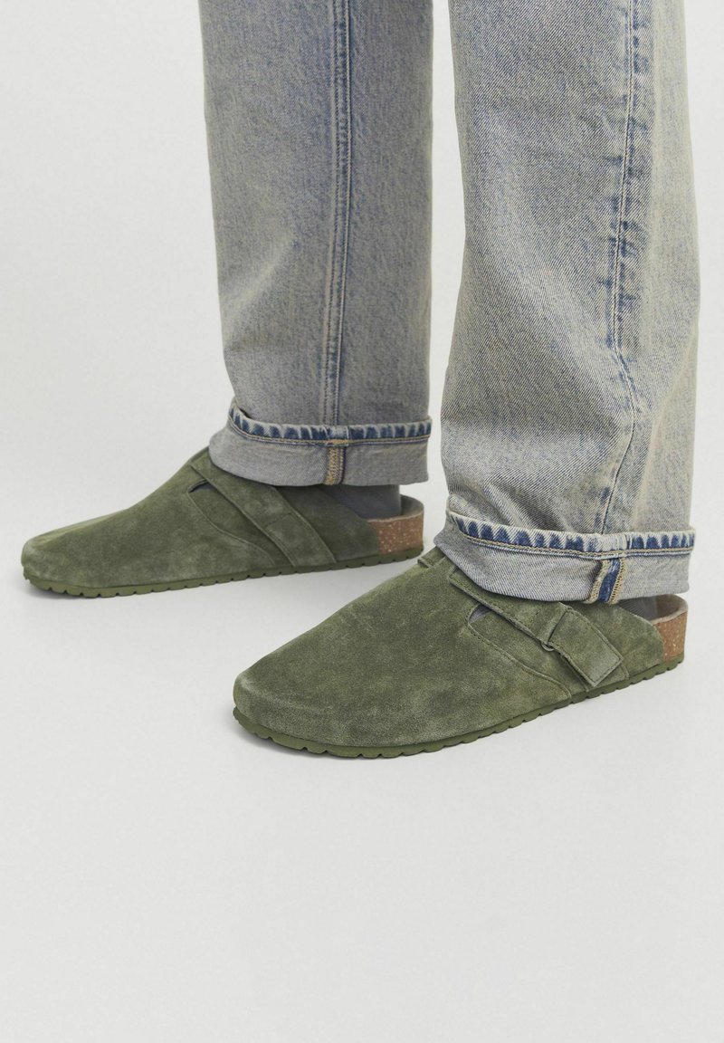 Zapatos de ante verde tipo slip-on con detalle de tira cruzada y acento de corcho en el talón, combinados con unos jeans de color azul claro, ligeramente arremangados.