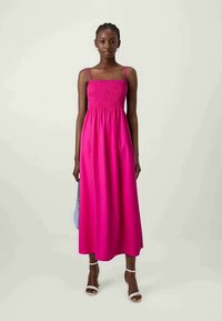 Fuksia lang kjole uten ermer med smocked liv, flytende skjørt og tynne stropper. Kombinert med hvite sandaler og en lyseblå veske.