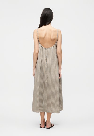 Γυναίκα με μακριά σκούρα μαλλιά, που φοράει ένα μπεζ και άσπρο ριγέ φόρεμα χωρίς μανίκια, με ανοιχτή πλάτη και λεπτές τιράντες, στέκεται σε απλό λευκό φόντο.