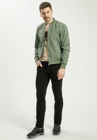 Jasnozielona kurtka bomberka, zapinana na zamek, z prążkowanymi mankietami, noszona na koszulce z grafiką, w zestawieniu z czarnymi obcisłymi dżinsami i ciemnymi sneakersami.