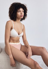 Balta bēru josta un rozā apakšbikses ar mežģīņu apdari, izgatavotas no mīksta materiāla. Modelis sēž pret neitrālu fona.