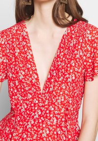 Vestido floral rojo con un escote en V profundo, mangas cortas abullonadas y una cintura ajustada con detalles de botones. La tela es ligera y texturizada.