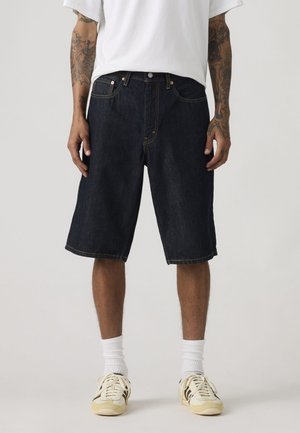 Mand iført mørke denimknælange shorts, hvide crew-sokker, beige sneakers, hvid T-shirt med synlige tatoverede underarme.