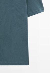 Texture de tissu de polo à manches courtes couleur sarcelle avec coutures visibles sur la manche et l'ourlet sur un fond blanc.