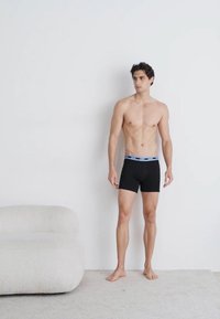 Boxer shorts de algodón negros con una cinturilla de color azul claro, diseño ajustado y textura suave, llevados por un modelo de pie sobre una alfombra clara.