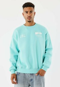 Homme avec des cheveux courts et bouclés portant un sweat-shirt bleu clair avec des logos blancs et un jean bleu délavé, se tenant contre un fond blanc uni.