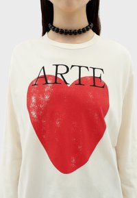 Crème-kleurig shirt met lange mouwen, voorzien van een versleten rode hartafbeelding en zwarte tekst "ARTE." Heeft een ronde halslijn en een zachte textuur.