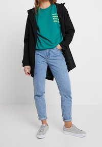 Giacca nera impermeabile sopra una t-shirt teal con testo bianco, abbinata a jeans azzurri chiari e sneakers grigie, caratterizzata da una vestibilità rilassata.