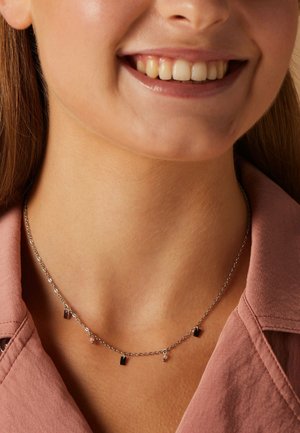 Zilveren ketting met kleine rechthoekige bedels en ronde kralen, tegen een huidkleurige achtergrond, gedragen met een roze blouse met kraag.