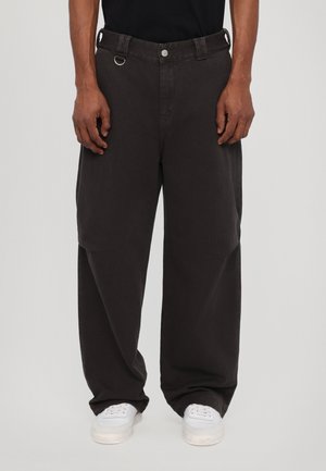 Hombre que lleva pantalones negros holgados con un botón plateado y detalle de lazo, combinados con zapatillas blancas y una camisa negra.