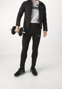 Zwarte sportieve trainingsoutfit met een rits-hoodie, strakke broek en een grijs T-shirt. Houdt dumbbells vast. Sneakers met groene accenten.