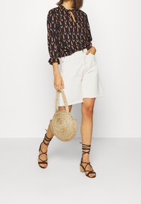 Blusa nera con motivo rosso, pantaloni corti di jeans bianchi, borsa di paglia intrecciata e sandali neri con cinturini; outfit casual con texture miste.