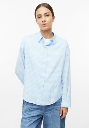 Woman with brown hair wearing a light blue checkered button-up shirt over a white top and blue jeans, standing with one hand behind her back.