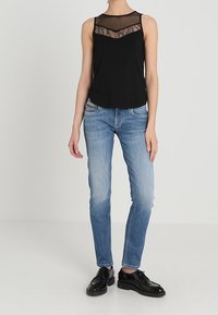 Top negro sin mangas con panel de encaje transparente, combinado con jeans de denim azul claro. La modelo lleva zapatos negros. Diseño simple y limpio.