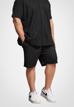Homme portant une chemise noire texturée boutonnée et un short assorti avec des baskets blanches Nike, mains dans les poches, debout sur un fond uni.