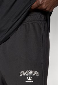 Μαύρο αθλητικό παντελόνι με ελαστική μέση, που φέρει λευκό λογότυπο "CHAMPION". Το υλικό φα ίνεται λείο, με ορατές πλαϊνές τσέπες.
