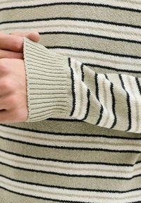 Mains ajustant le poignet d'un pull beige à rayures horizontales noires et blanches avec des poignets en maille côtelée.