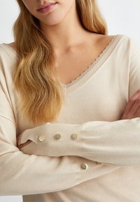Maglione beige con scollatura a V e bordo ondulato. Presenta maniche lunghe con accenti di bottoni dorati vicino ai polsini. Texture morbida.
