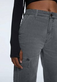 Graue Cargo-Hosen mit glatter Textur, Seitentaschen und einem Knopfverschluss am Bund. Kombiniert mit einem schwarzen Langarmoberteil.