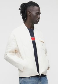 Baltas siūtas bombonešis su priekyje esančiu užtrauktuku, viena krūtinės kišenė ir ribotu kaklu, dėvimas ant tamsiai mėlynos dryžuotos marškinėlių su raudonu akcentu.