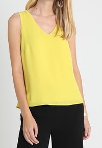 Femme portant un chemisier jaune sans manches à col en V, un pantalon noir et des boucles d'oreilles en forme de créoles dorées, sur un fond blanc uni.