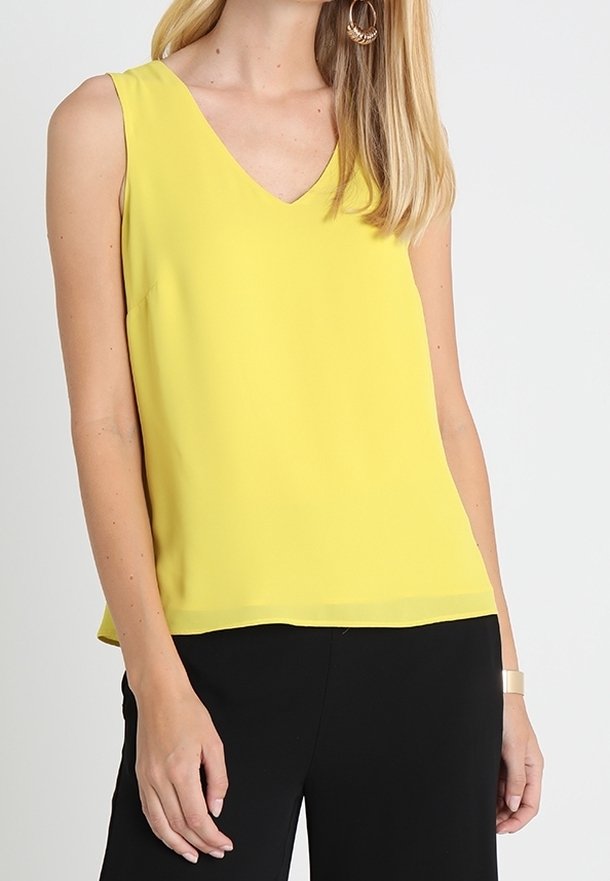 Femme portant un chemisier jaune sans manches à col en V, un pantalon noir et des boucles d'oreilles en forme de créoles dorées, sur un fond blanc uni.