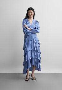 Frau steht mit verschränkten Armen, trägt ein langes, geschichtetes blaues Kleid und schwarze offene High Heels vor einem einfarbigen grauen Hintergrund.