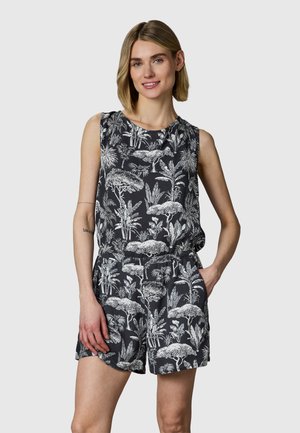 Kobieta z krótkimi blond włosami, ubrana w czarny bezrękawnik z białymi tropikalnymi nadrukami drzew i ptaków, ręka w kieszeni, uśmiechnięta.