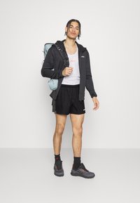 Pantalones cortos deportivos negros, camiseta sin mangas de malla blanca, chaqueta oscura con cremallera, zapatillas negras con acentos naranjas, mochila azul claro, pose de pie.
