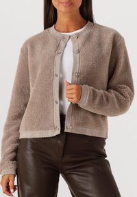 Bruine fleecejas met een ronde halslijn, knoopsluiting en geribbelde afwerking. Heeft een zachte textuur en een kort model.