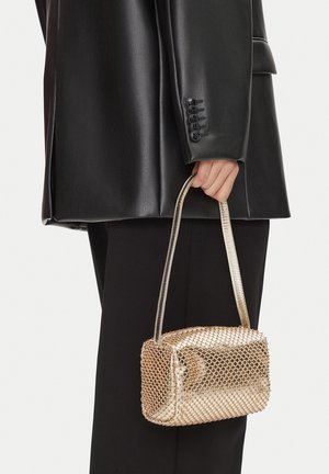 Mano che tiene una piccola borsa a rete dorata con tracolla sottile, indossando una giacca di pelle nera e pantaloni neri, vista dalla vita in giù.