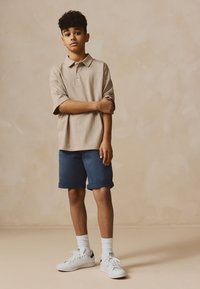 Polo beige, pantalones cortos azul marino con puños, zapatillas blancas y calcetines blancos. Corte casual, tela suave, diseño sencillo y sin patrones visibles.