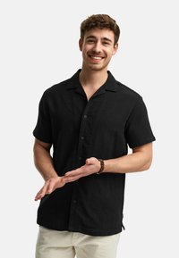 Short-sleeve, black button-up shirt made of textured fabric with a folded collar and contrasting buttons, paired with light-colored pants.