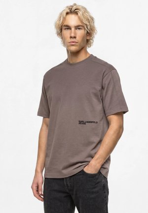 Homme blond portant un t-shirt taupe Karl Lagerfeld Jeans à manches courtes et un jean noir, debout avec une main dans la poche.