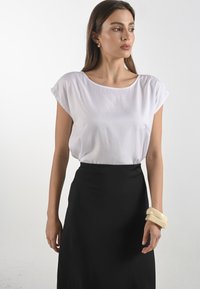 Blusa bianca a maniche corte abbinata a una gonna nera. La modella indossa bracciali sovrapposti di colore crema su un polso, evidenziando una texture liscia.