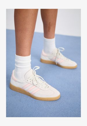 Witte en roze suède sneakers met drie roze strepen, een lichtbruine rubberen buitenzool en witte geribbelde sokken. Geplaatst op een blauw tapijt.