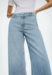 Jeans de mezclilla de tiro alto en lavado claro y pernera amplia, con cierre de botón frontal y una textura sutil. Sin patrones ni acentos visibles.