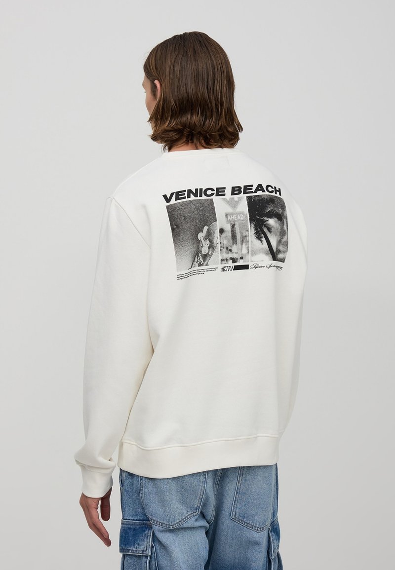 Kremowy sweter z okrągłym dekoltem, z grafiką zawierającą napis "VENICE BEACH" oraz obrazami na plecach, wykonany z miękkiego materiału.