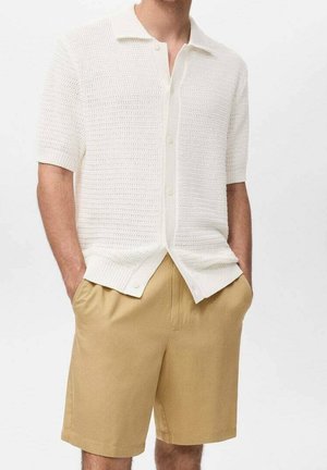Mand iført hvid kortærmet strikket skjorte med knapper og beige knælange shorts med hænder i lommerne.