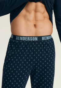 Męski brzuch z widocznymi mięśniami brzucha, podnoszący ciemną koszulę, w błękitnych spodniach od pidżamy z małymi wzorami tipi w kolorze białym oraz pasem w kolorze "HENDERSON".