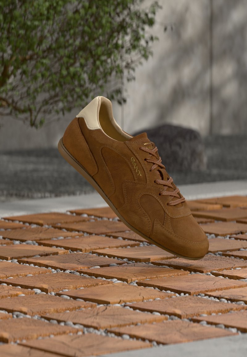 Baskets en daim marron avec col de talon beige et logo, lacets noués, flottant au-dessus d'un sol en bois et pierre à motifs en plein air.