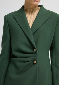 Vihreä räällä valmistettu bleiseri, jossa on terävä kaulus, sivurypytys ja kolme koristeellista kultanappia. Kangas näyttää olevan teksturoitu ja jämäkkä.