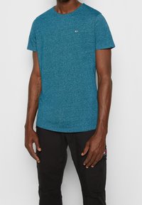 Tyrkysové melírované tričko s kulatým výstřihem, vyrobené z měkkého materiálu, s krátkými rukávy a malým logem na hrudi. V kombinaci s černými cargo kalhotami.