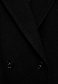 Manteau noir à double boutonnage avec une texture douce, présentant des revers proéminents et des boutons noirs. Lignes épurées et coupe ajustée.