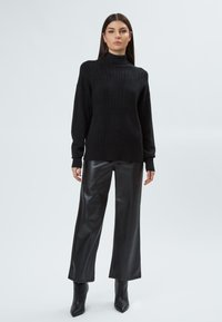 Schwarzer Strickpullover mit hohem Kragen und Zopfmuster, kombiniert mit schwarzen Leder-Hosen mit weitem Bein und spitzen Knöchelstiefeln.